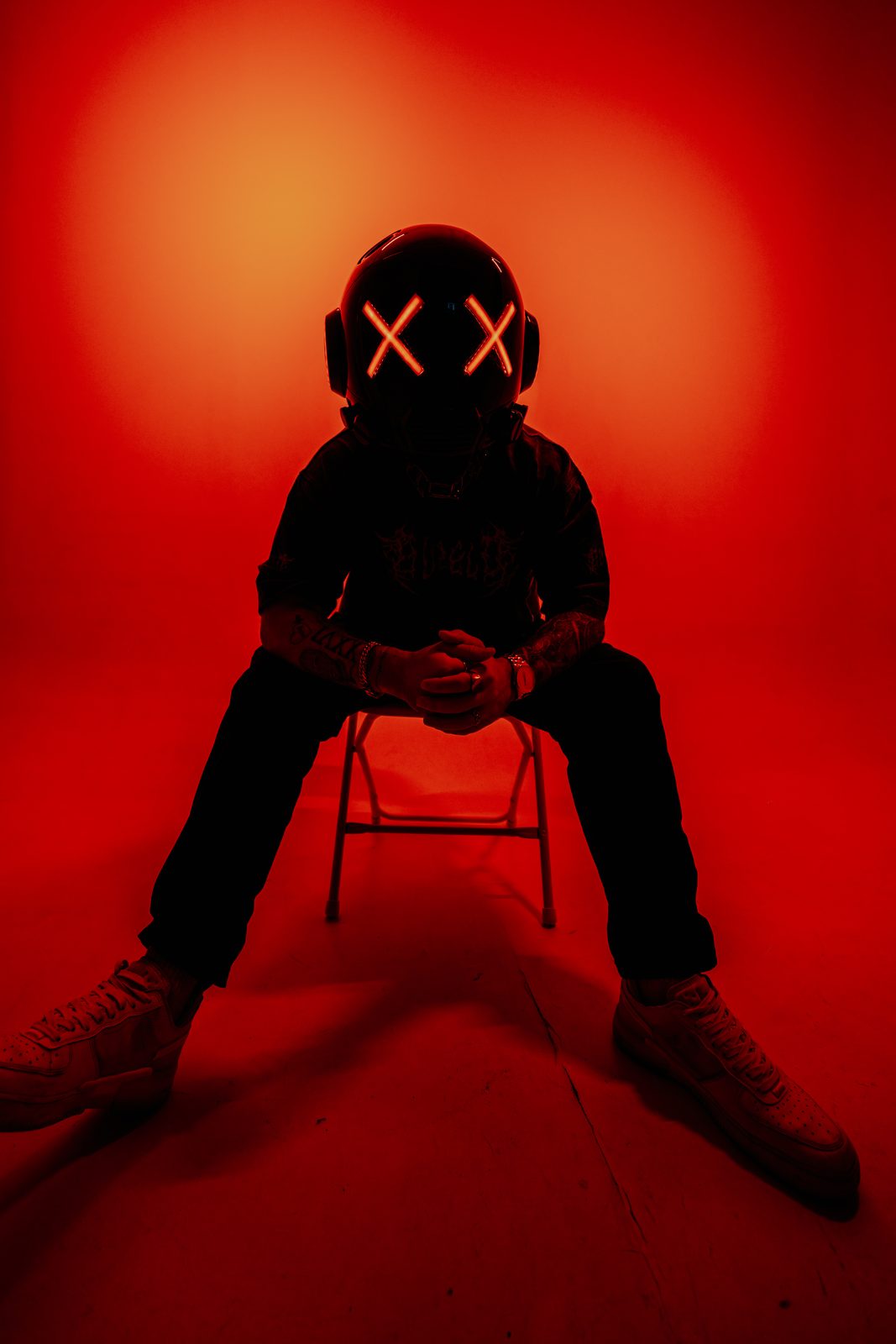 Helmeted DJ seated on a folding chair in red light, legs wide
